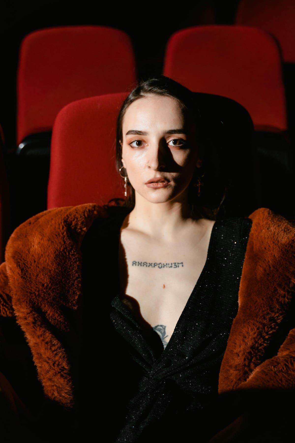 A young woman seated alone among rich red theatre seats, lit warmly.